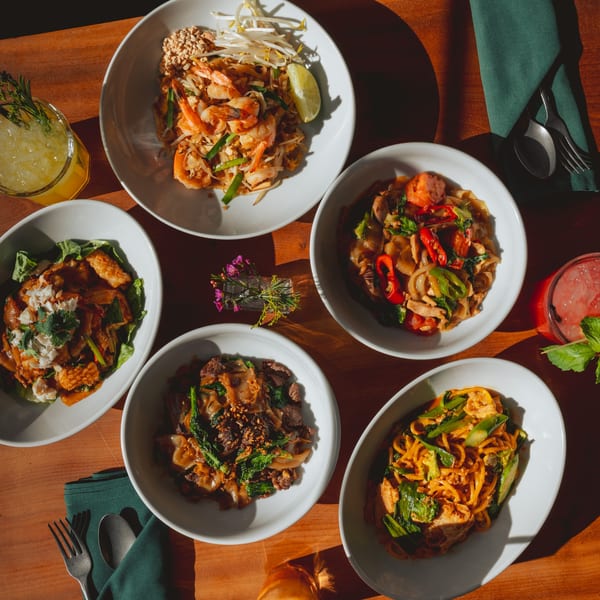 Overhead view of five Thai noodle dishes and two cocktails on a wooden table.