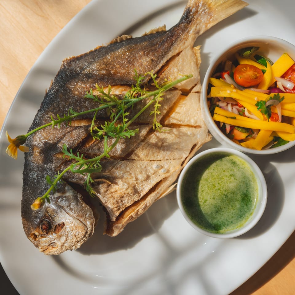 Crispy fried whole fish with mango salad and green dipping sauce on a white plate.