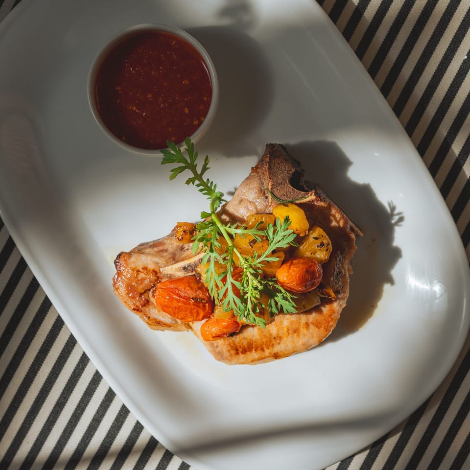 Grilled pork chop with roasted tomatoes, pineapple, and fresh herbs, served with a side of sauce.