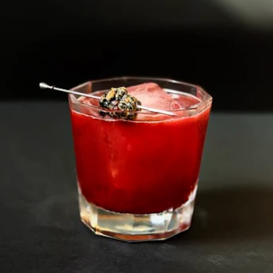 A vibrant red cocktail in a faceted glass, garnished with a blackberry skewer, against a dark background.