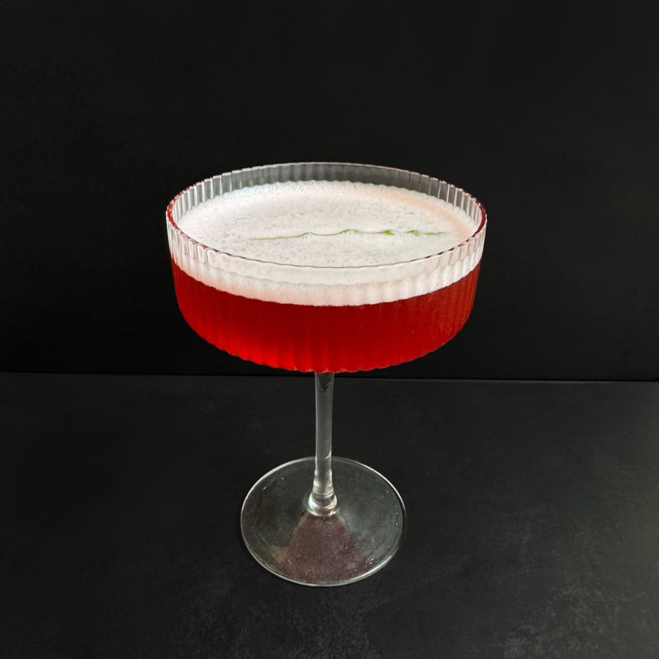 A vibrant red cocktail with a frothy white top, garnished with a green leaf, in a coupe glass.