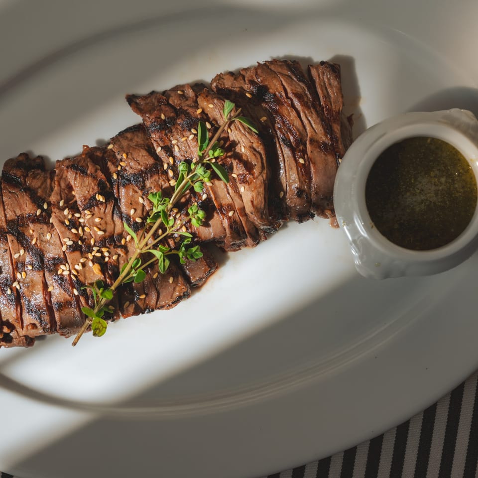 Grilled sliced steak with sesame seeds and a sprig of thyme, served with green sauce.