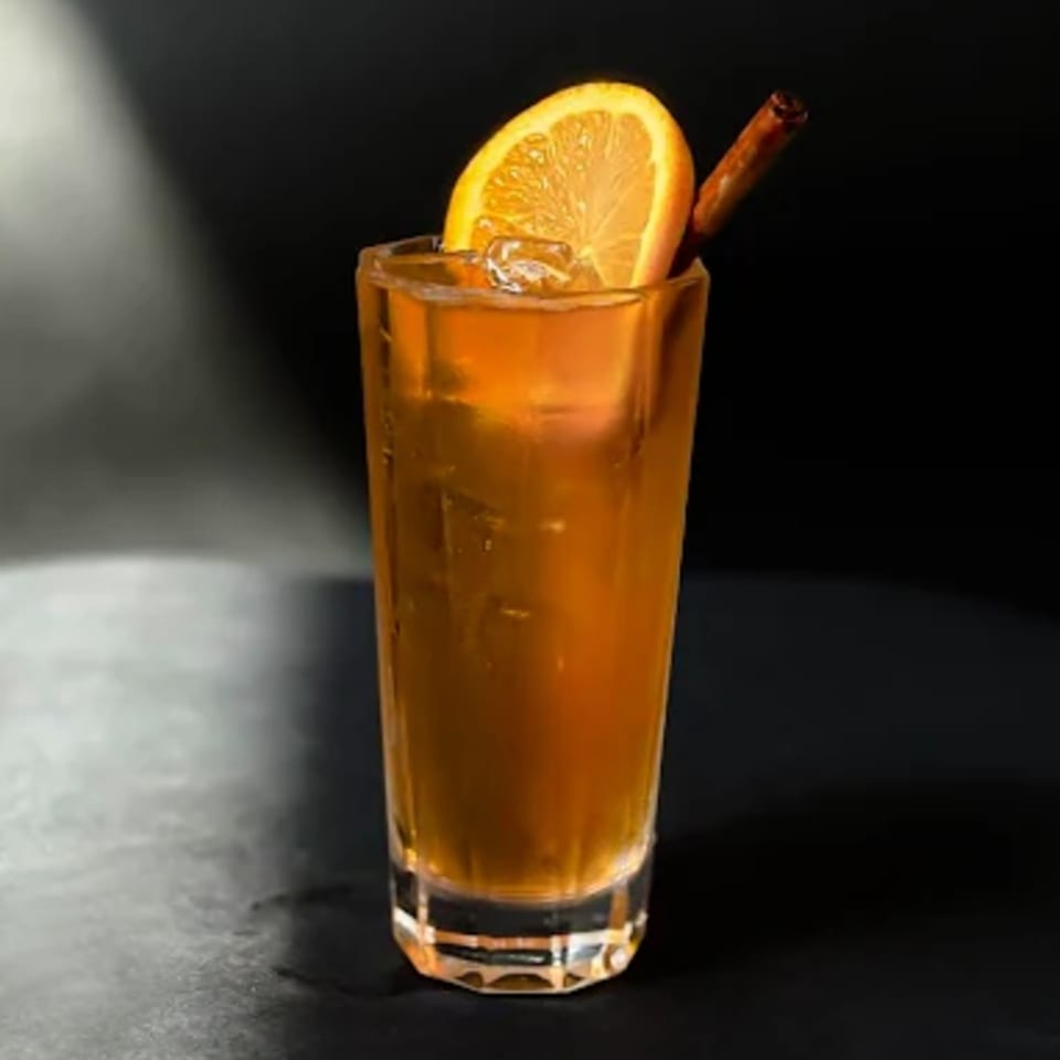 A tall glass of iced tea with an orange slice and cinnamon stick on a dark surface.
