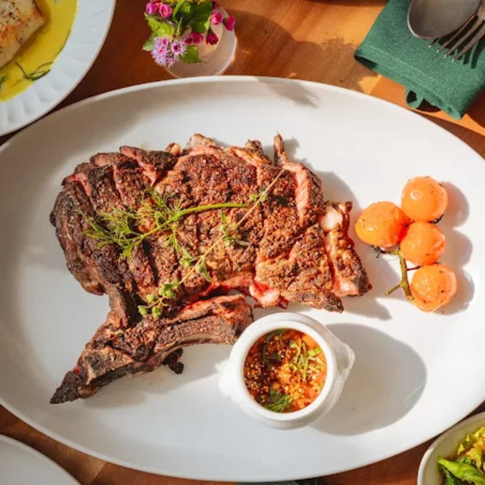 Grilled steak with herbs, cherry tomatoes, and a side of sauce on a white plate.