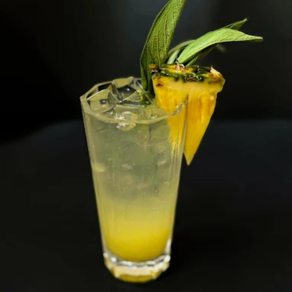 A tall glass of iced yellow cocktail, garnished with pineapple and green leaves, against a dark background.
