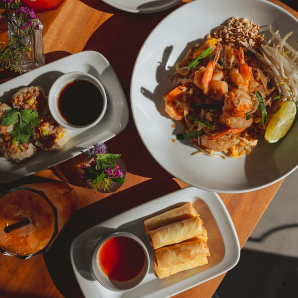 Overhead view of Teakwood Thai Chophouse dishes: shrimp pad thai, dumplings, spring rolls, and Thai iced tea.